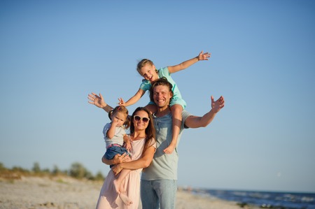 Young happy family on the beach. A woman is holding a younger daughter in her arms. The second girl sits on her father's shoulders. Joy of communication with children and nature.の写真素材