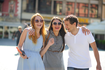 Three young people stand on the square of the ancient city. They are looking at beautiful buildings with pleasure. Friends are hugging each other. Everyone is in a good mood.の写真素材