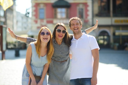 Three young people stand on the square of the ancient city. They are looking at beautiful buildings with pleasure. Friends are hugging each other. Everyone is in a good mood.の写真素材