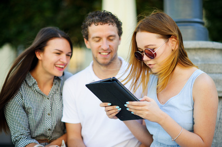 Two girls and the guy with interest look at the tablet screen. Friends cheerfully communicate.の写真素材