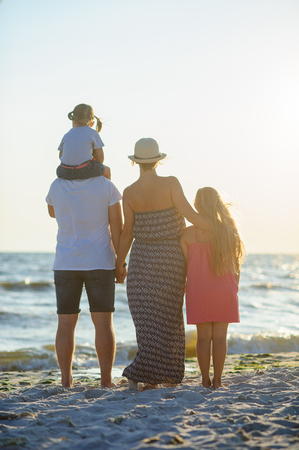 Father, mother and two daughters standing on the sand and admiring the sea. Younger girl sitting on the shoulders of his father. The blue sky, the sun, fresh sea wind. Rear view.の写真素材