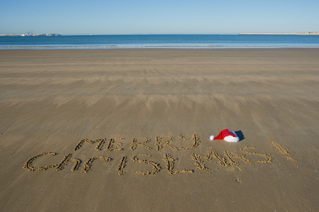 Merry Christmas! Someone has written a congratulation on sea sand. A bright cap of Santa Claus on brown sand. Sun, sea, blue sky. Christmas in the warm regions.の写真素材