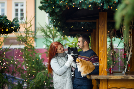 Christmas holidays. Young couple on the Christmas bazaar with pets. Two little spitz-dogs on hands at the owners.の写真素材