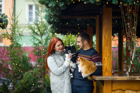 Christmas holidays. Young couple on the Christmas bazaar with pets. Two little spitz-dogs on hands at the owners.の写真素材