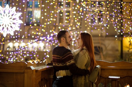 Young couple stands embracing against the shining festive garlands. Lovers look at each other with tenderness. Night. Romantic rendezvous.の写真素材