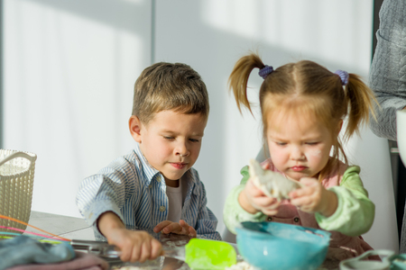 Two small children prepare something from the dough. A boy and a girl are sitting at the kitchen table. Their faces are stained with flour. Children try hard.の写真素材