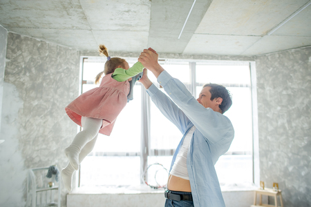 Dad plays with his little daughter. The man lifts the baby by the handles. In his eyes, love and tenderness. Against the background of a large window.の写真素材
