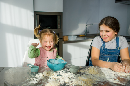 Two little girls are cooking something in the kitchen. The children are sitting at the table. They knead the dough. Hands and faces of girls in flour.の写真素材