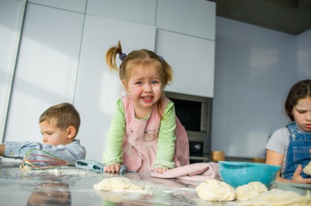 Three children prepare something from the dough. Two sisters and a brother are sitting at the kitchen table. There are various kitchen utensils on the table. Children like to cook food. All are stained with flour.の写真素材