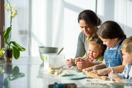 Mother teaches her three children to cook. Family is preparing something from the dough. There are necessary products on the kitchen table. Useful and fun pastime.の写真素材