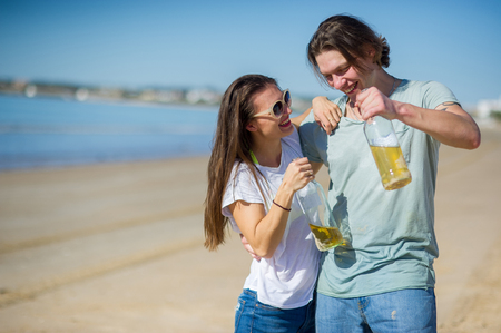 Cheerful young couple drinks wine on the beach. In hands at young people glass bottles with drink. They have an excellent mood.の写真素材