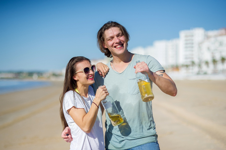 Cheerful young couple drinks wine on the beach. In hands at young people glass bottles with drink. They have an excellent mood.の写真素材