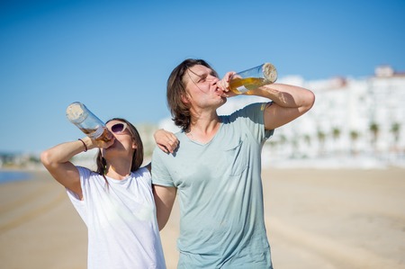 Cheerful young couple drinks wine on the beach. In hands at young people glass bottles with drink. They have an excellent mood.の写真素材