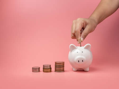 White piggy Bank and coin and human hand putting coin in piggy bank on a pink background. Copy space for design.の写真素材