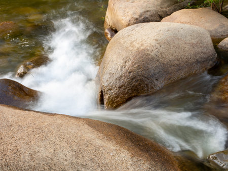 A long-exposure shot of a small creek flowing through a brown rock. And there are green trees as a...