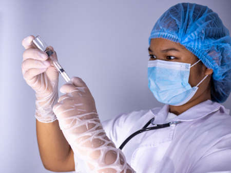 Studio portrait of a female doctor wearing a mask and wearing a hat. In hand was a bottle of vaccine and a sling of syringes. standing on a white background. Studio shot background, COVID-19 conceptの写真素材