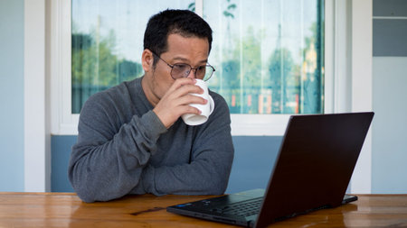 Asian men wear long sleeves and eyeglasses.He is drink a coffee and Sitting at home working on a...