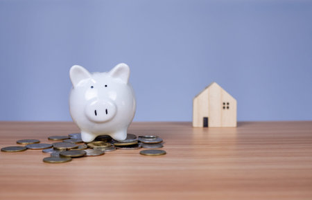 A white piggy bank is placed on a pile of coins. And beside it is a wooden model house on a white...