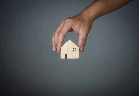Concept of selling a house. A hand is holding a model house on a gray background. Real estate agent offer house, property insurance and security, affordable housing concepts, home insurance broker agent, salesman person.の写真素材