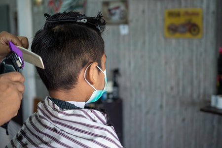 Teenage boy in a face protective mask is getting a haircut from a barbershop. Fashionable elongated...