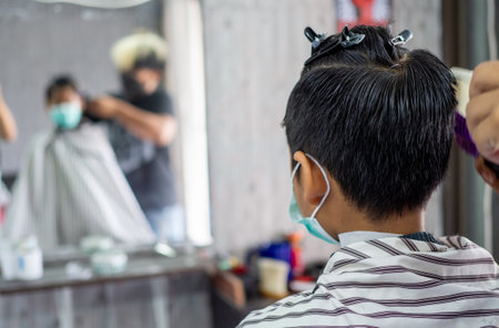 Teenage boy in a face protective mask is getting a haircut from a barbershop. Fashionable elongated haircuts for boys. Beauty salon in quarantine coronavirus covid-1の写真素材