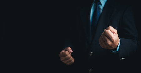 Businessman in a suit stands and makes a fist gesture on a dark background.の写真素材