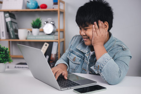 Young Asian boy smiling while using a laptop at his desk, representing successful online learning,...