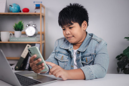 Asian boy using smartphone and laptop at home for online learning, digital communication, or entertainment. Smart kid in denim jacket.の写真素材