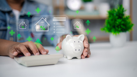 Financial planning concept with person using calculator beside piggy bank and digital overlay of home icons, percentage signs, and checklist. Mortgage, home loan, savings, real estate investment.の写真素材