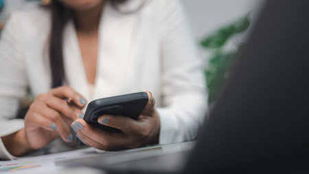 Businesswoman holding smartphone while working with financial charts and laptop on office desk....