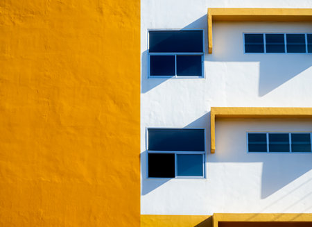 Modern yellow and white building facade with blue glass windows and shadow, minimal architecture background, abstract geometric design with sunlight contrast.の写真素材
