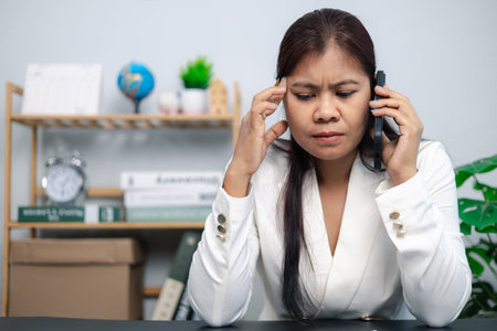 Businesswoman feeling stressed and frustrated while talking on mobile phone at office desk, concept...
