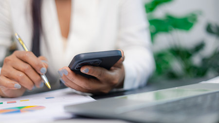 Businesswoman using smartphone and analyzing financial report at office desk, representing multitasking, business management, and digital productivity concept.の写真素材