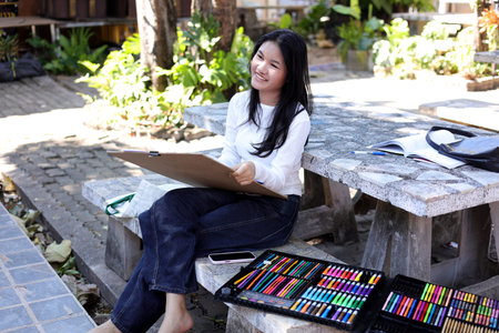 Asian teenage girl drawing outdoors with colorful art supplies, creativity activity, student...