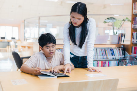 An older Asian girl helps a young boy read a book in a bright library. Concept of education,...