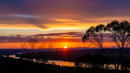 A sunset over a lake with trees and mountains in the background. The sky is painted with hues of orange and purple, with clouds scattered across it. The trees are silhouetted against the vibrant sky, adding depth to the scene.の素材
