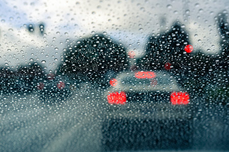 Raindrops on the windshield on a rainy day; blurred car stopped at a traffic light in the...