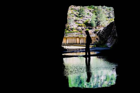 Man sitting in front of the exit to a tunnel, silhouette reflected in a shallow pond; Hetch Hetchy reservoir area, Yosemite National Park, Sierra Nevada mountains, Californiaの写真素材