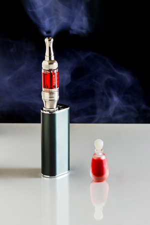 A black Electronic cigarette (or e-cigarette) emitting vapor and a small bottle full of red e-liquid on dark background.の写真素材