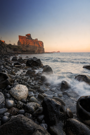 Italian destination: Acicastello in Sicily at sunset against a clear blue sky. Photo taken by the beach; waves on foreground.のeditorial素材