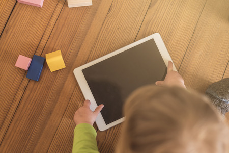 top view of a toddler playing with a tablet device on a wooden floor with toys in the backgroundの写真素材