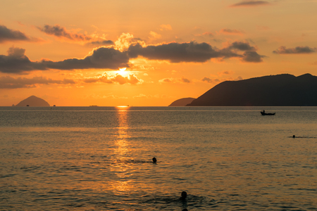 Orange trace of sunlight on water of sea at dawn with clouds and swimming people in waterの写真素材