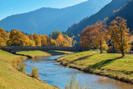 Wooden bridge over a calm river in an autumn park with orange trees in the mountains in the Alpsの写真素材
