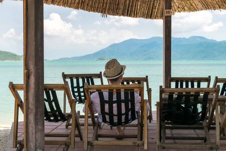 Young man with hat sits in chair on beach under canopy and look at sea on vacationの写真素材