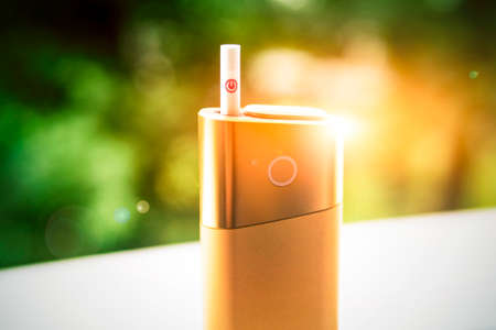 A new kind of electronic cigarette. IQOS tobacco heating system. Mod for smoking stands with a cigarette. Heated cigarettes for smoking. Safe form of smoking. Against the background of green treesの写真素材