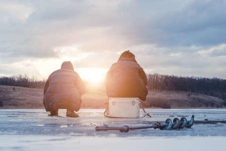 Men fishing on ice, winter fishing on a frozen river in the evening at sunsetの写真素材