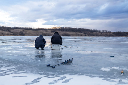 Men fishing on ice, winter fishing on a frozen river in the evening at sunsetの写真素材