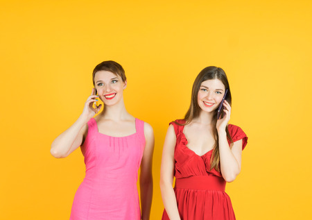Technology, friendship and leirure concept - two smiling young women talking with smartphones. Isolated studio, yellow background, female model.の写真素材