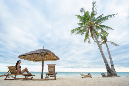 Young happy girl sitting on beach chair with umbrella and palm tree on the beachの写真素材