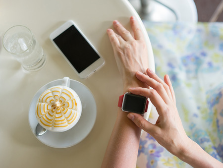 Womans hands with smartwatch, smartphone, coffee and glass of water on the table. Focus on smartwatch.の写真素材